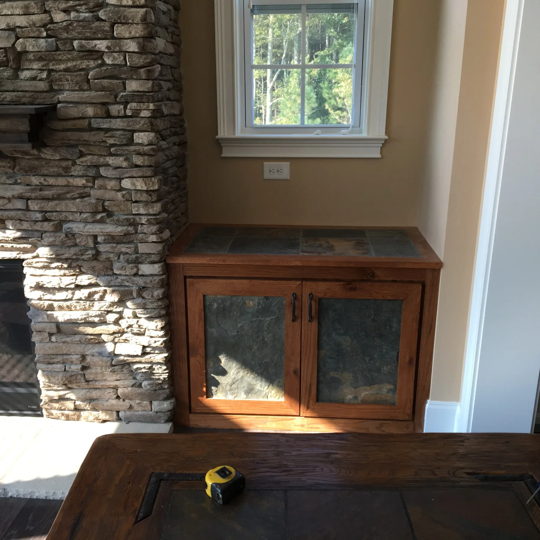 Oak cabinet with slate panel doors beside a stacked stone fireplace, tape measure on the bench